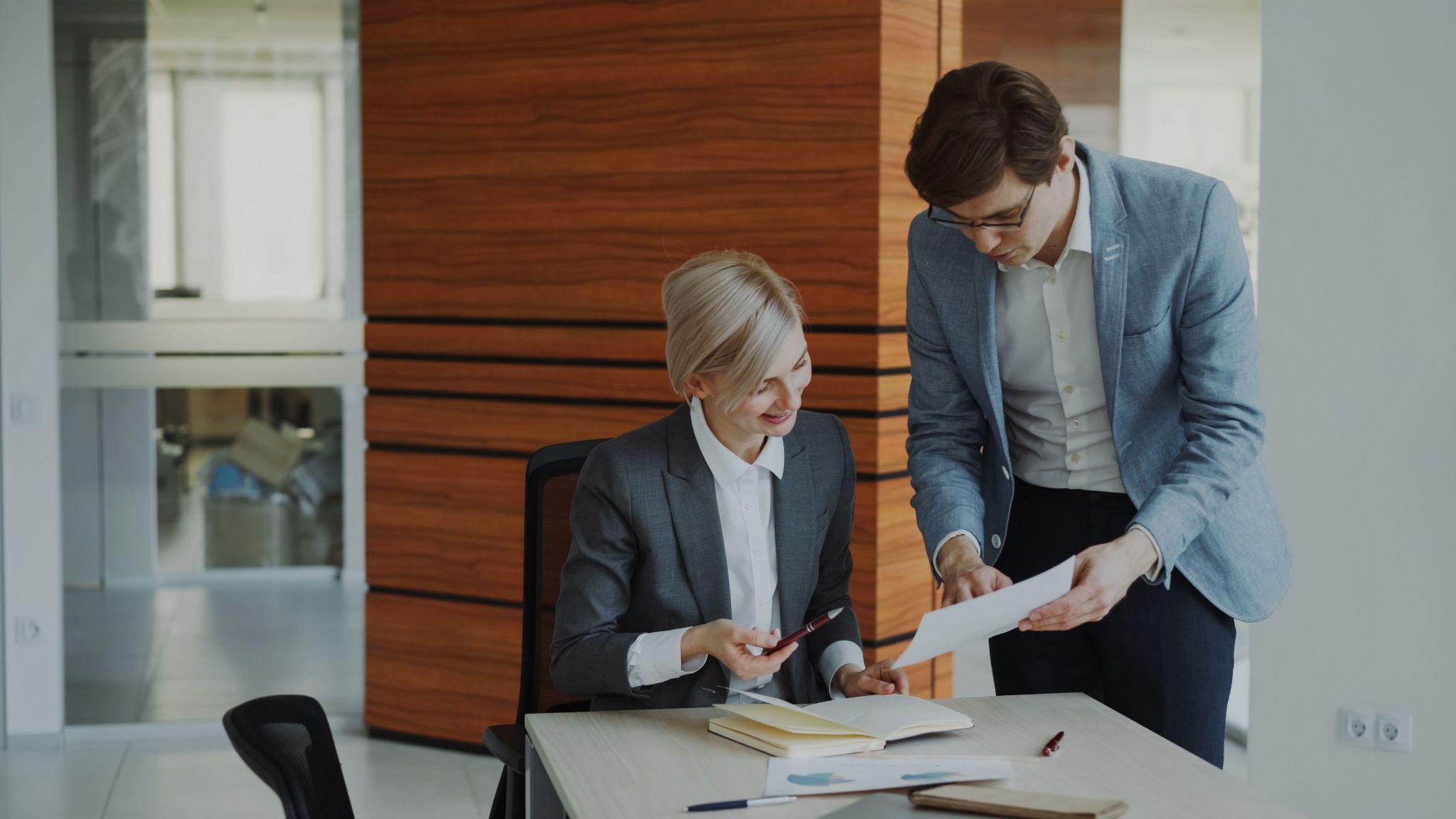 A businesswoman in a gray blazer reviews a document with a male colleague in a blue blazer standing beside her desk in a modern wood-paneled office.