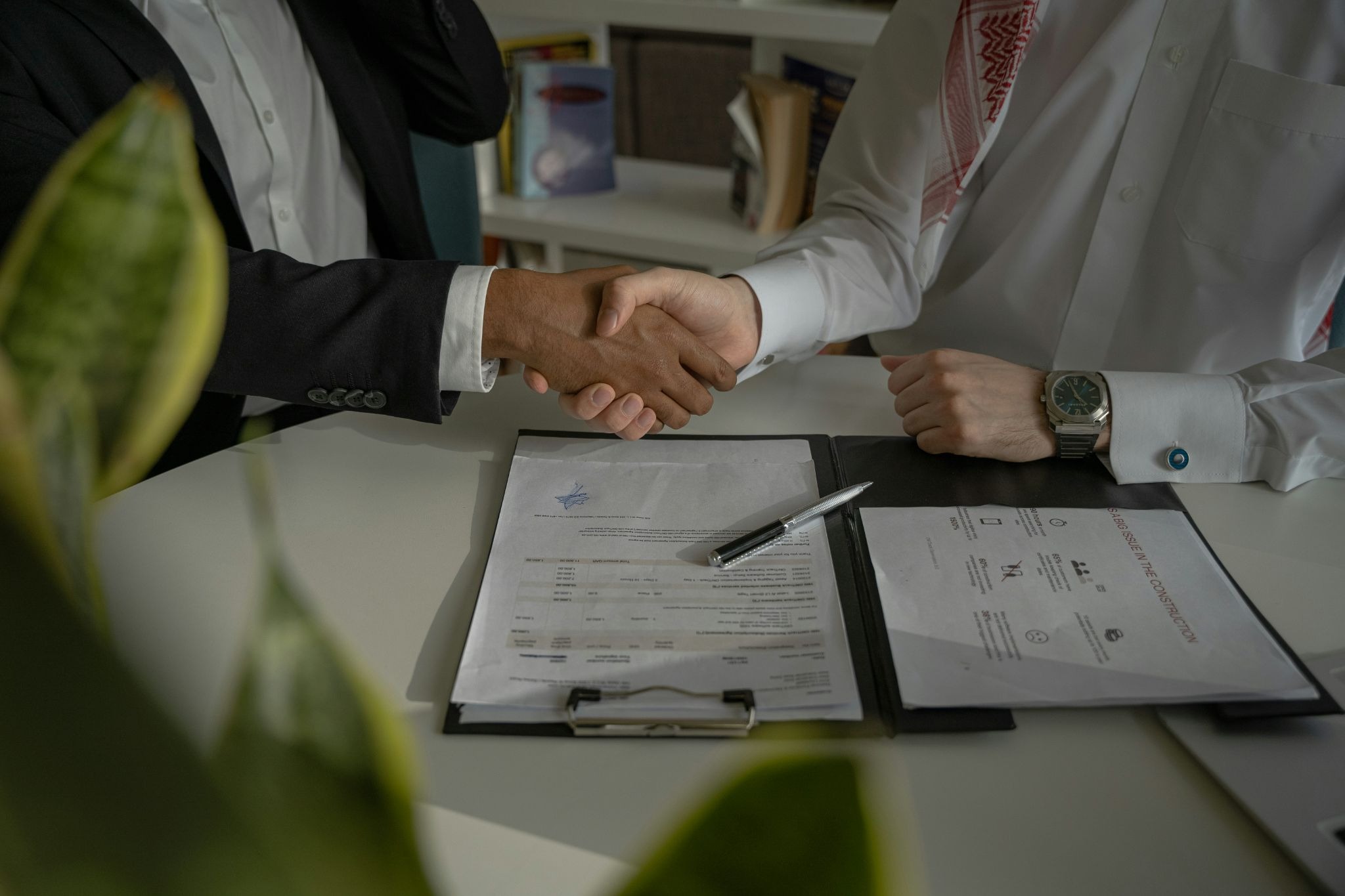 Two businesspeople shake hands over a desk with signed contract documents and clipboards, plants in the foreground.