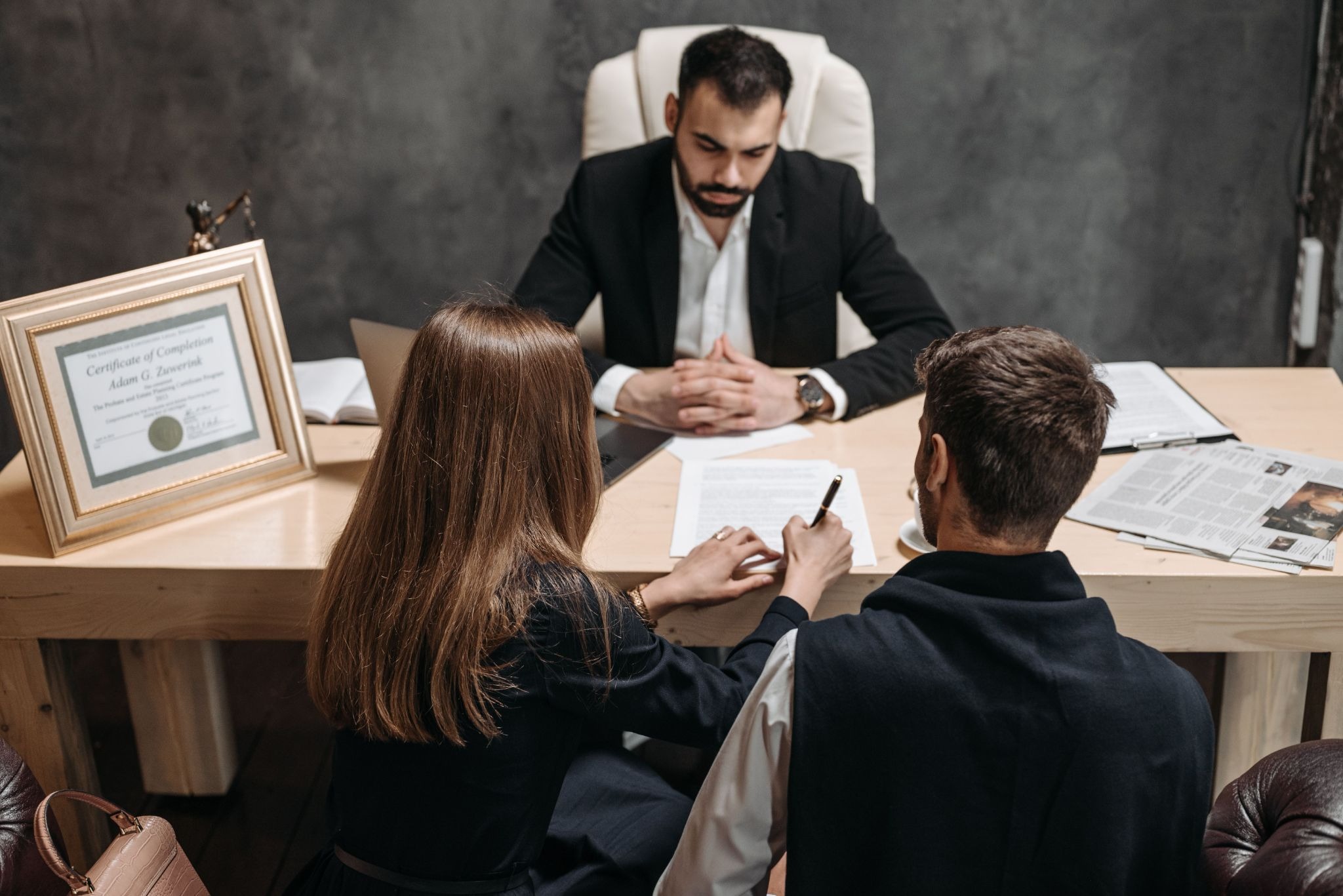 Two clients signing a document at a lawyer's desk, with a certificate of completion displayed on the desk.